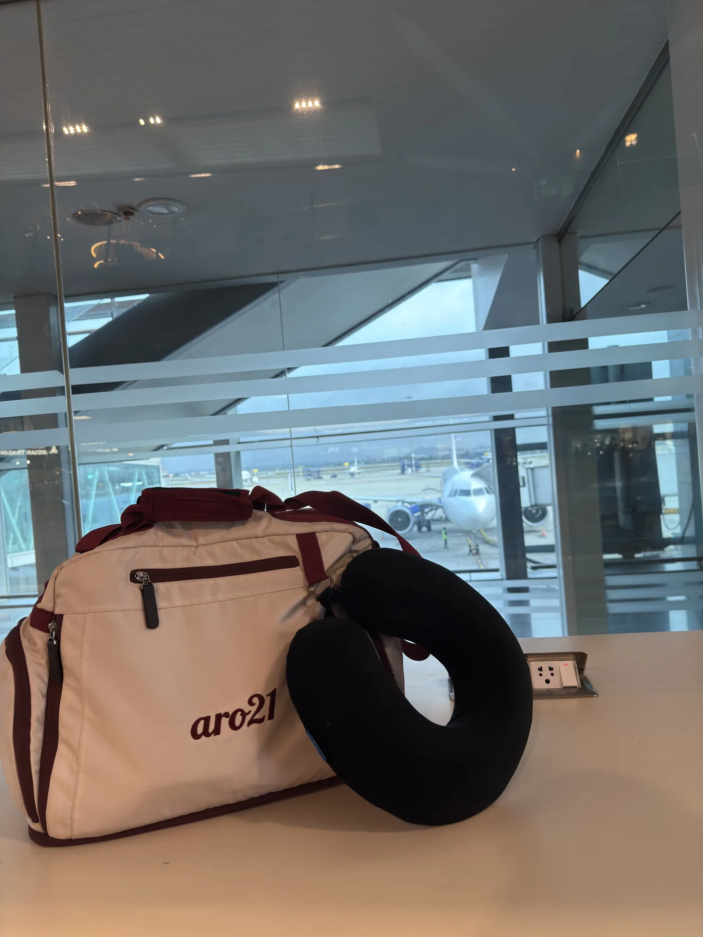 The Dawn Duffel resting at an airport gate with a travel pillow looped over the handle, an aircraft on the stand beyond the glass